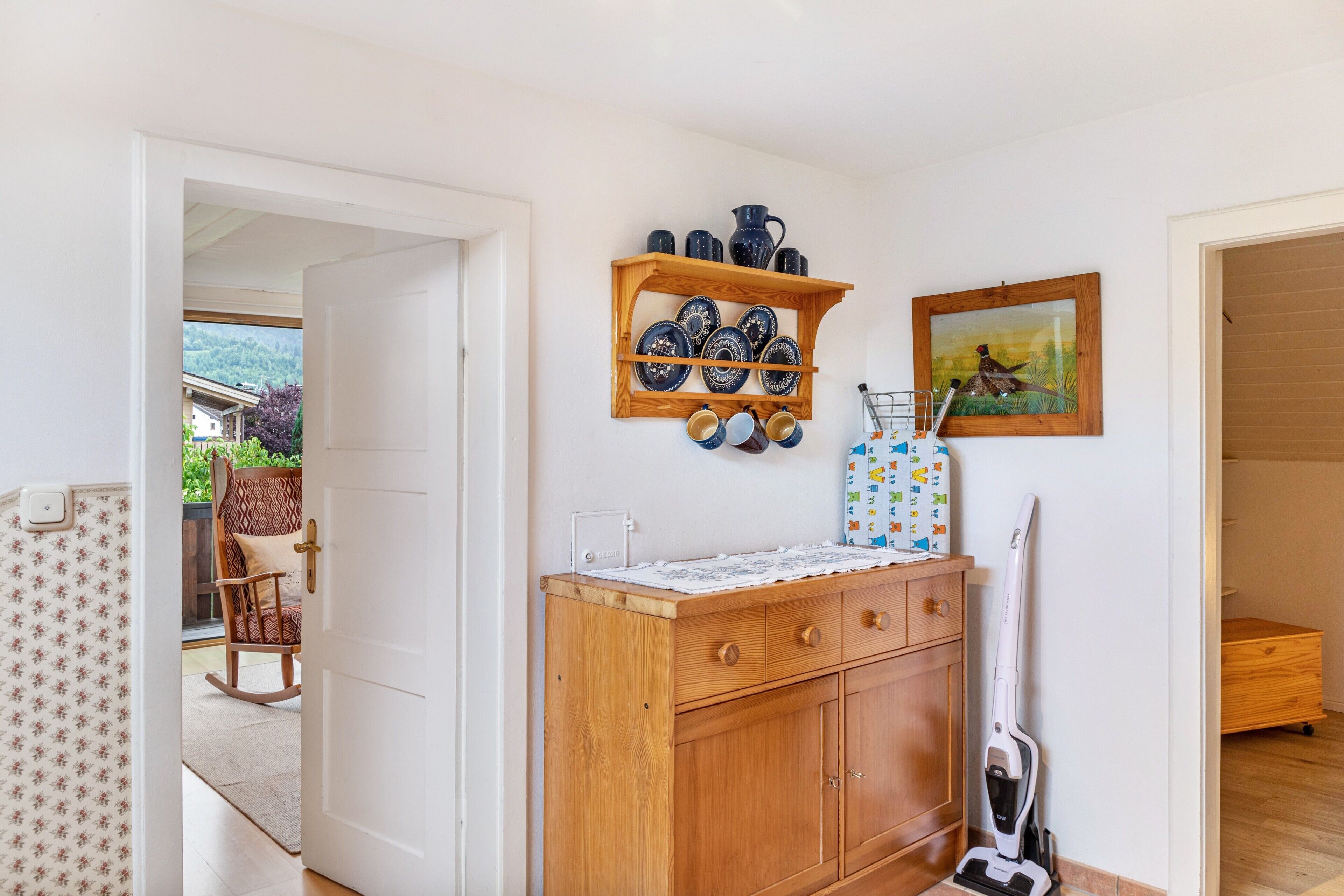 Een kamer met een houten kast, een wandplank met servies, een schilderij en een stofzuiger.