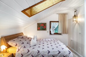 A bedroom with a wooden ceiling, a bed with a patterned bedspread, a nightstand with a lamp, a window with curtains, and a wall-mounted light fixture.