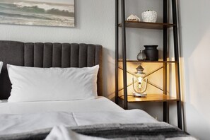 A bed with a dark headboard, white pillows, a bedside table with a lamp, and a shelf with decorative items.
