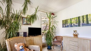 A room with a television, a large potted plant, a wicker chair with a teddy bear, a wooden cabinet, and a wall-mounted painting.