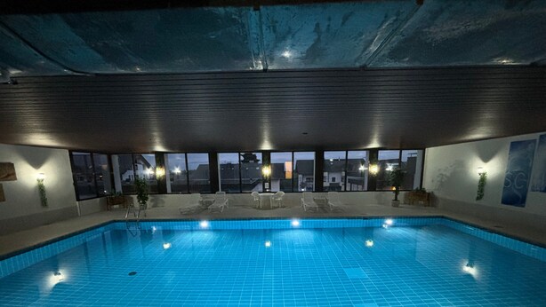 Una piscina interior con baldosas azules cristalinas, sillones de descanso y grandes ventanales que ofrecen vistas del paisaje urbano.