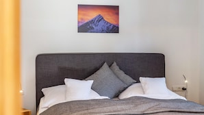 Een bed met een grijs hoofdbord, witte kussens en een schilderij van een berg aan de muur.