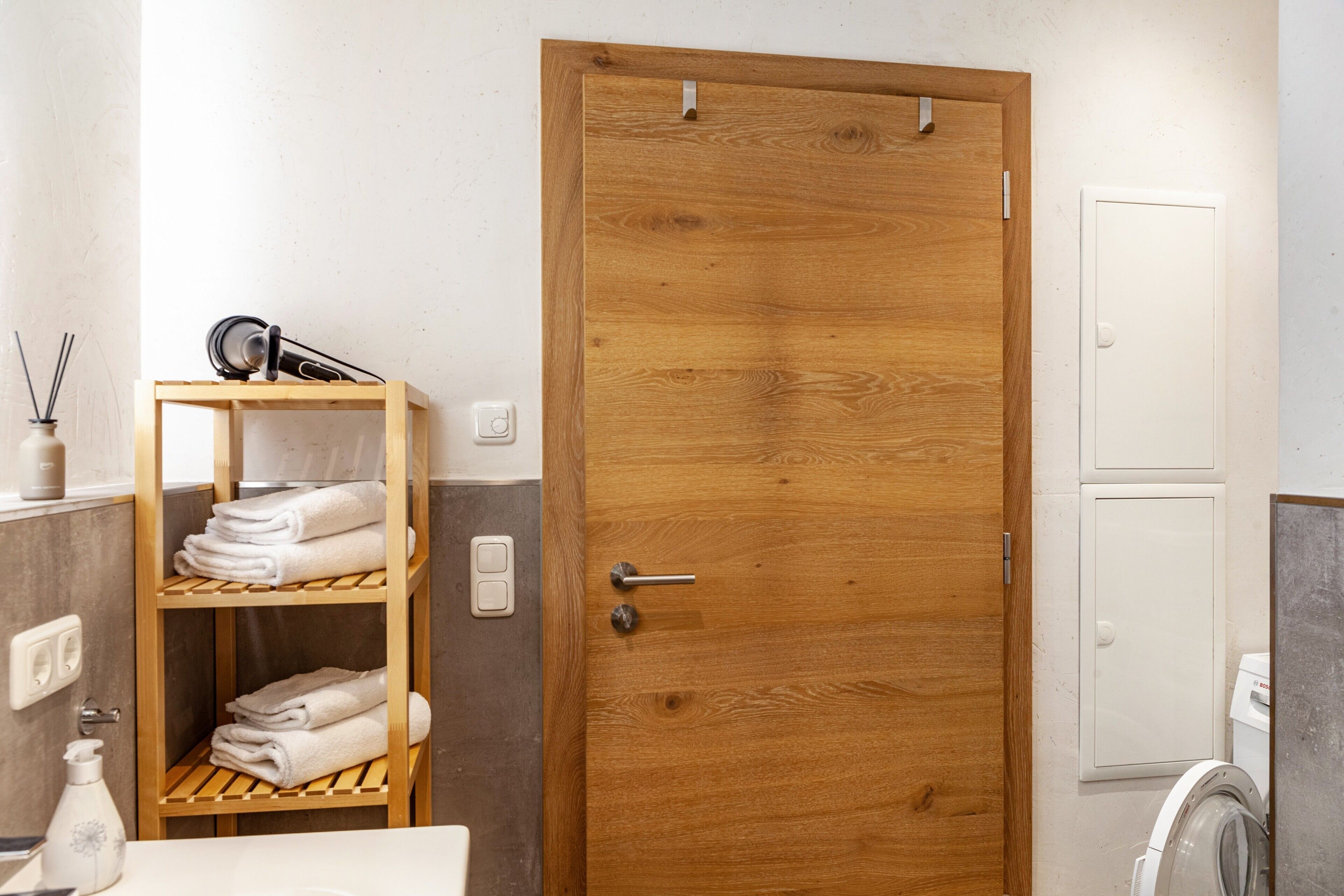 A wooden door with a handle, a shelf with folded towels, a white washing machine, and a small table with a bottle and a candle.
