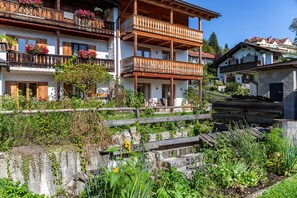 Un edificio a più piani con balconi, ringhiere in legno e un’area giardino con piante e fiori.