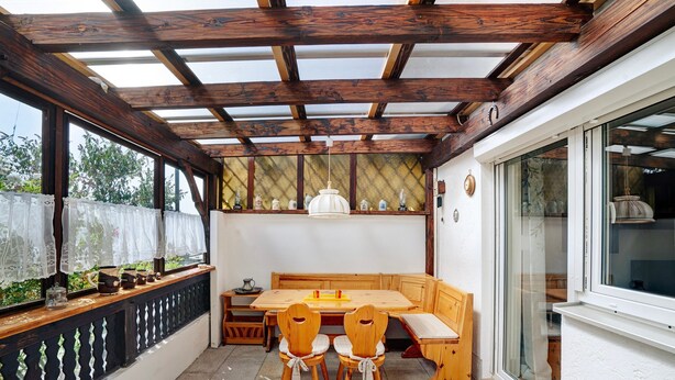 A covered patio with a wooden table and chairs, a bench, and a glass door leading to an interior space.