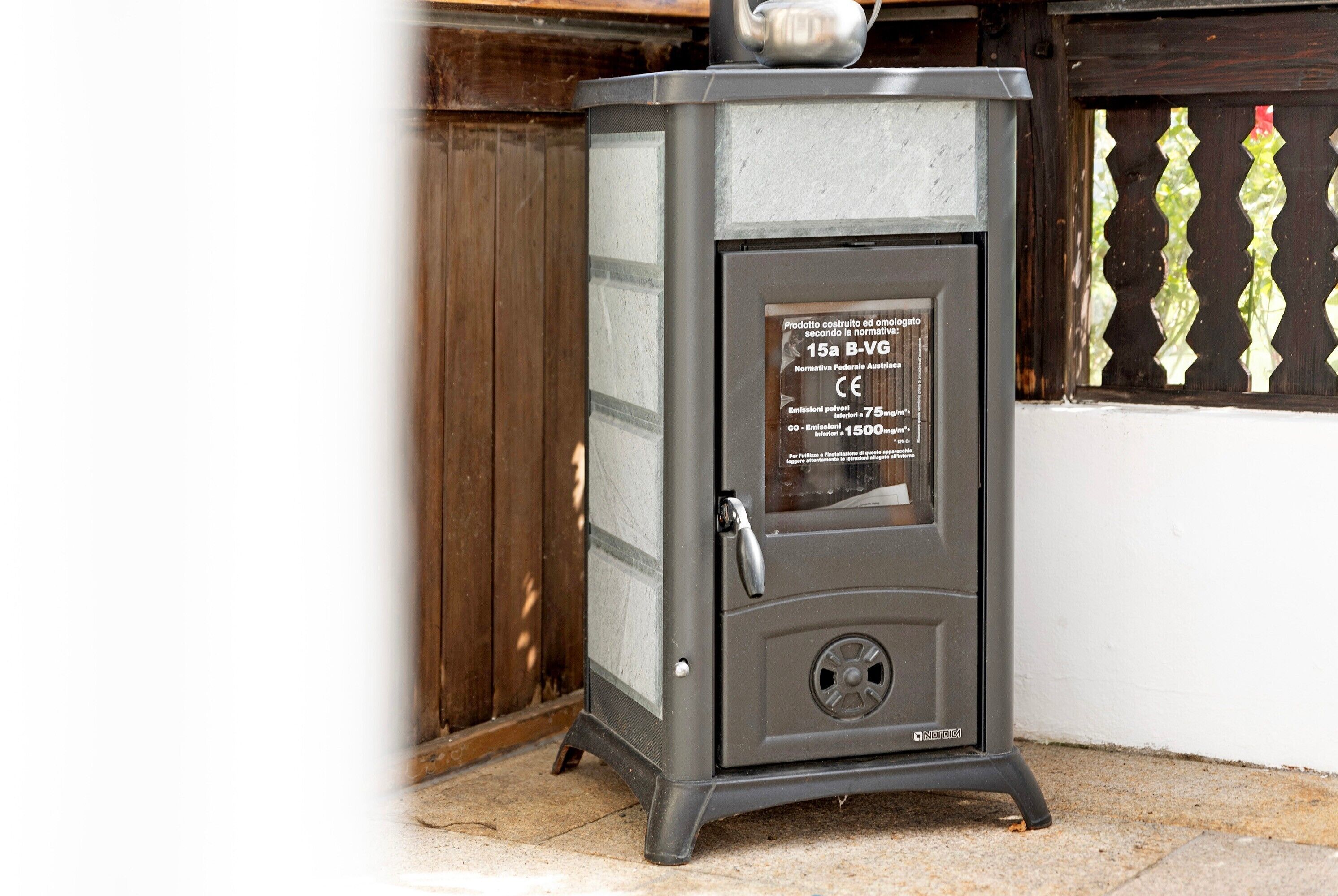 A gray, insulated wood stove with a CE certification label, positioned against a wooden wall and a window with decorative glass.