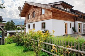 A two-story wooden house with a balcony, a fenced yard, and a mountain backdrop. - Apartment "Falkenstein" with Mountain View, Two Balconies, Private Sauna (Garmisch-Partenkirchen)