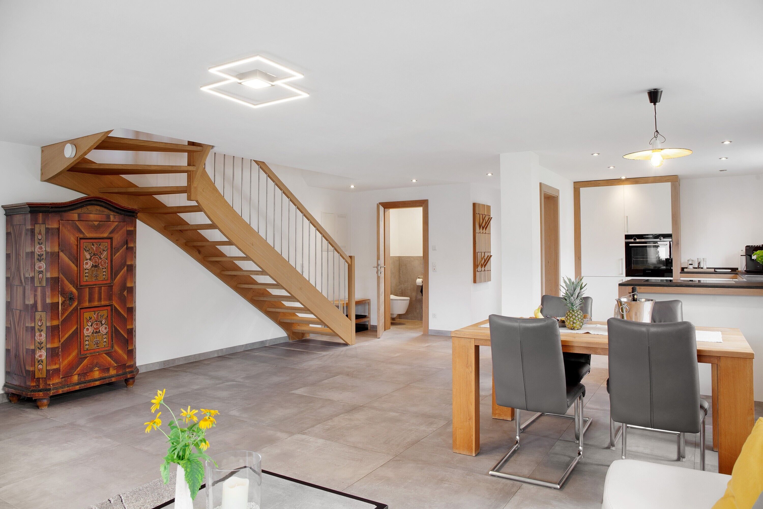 Un intérieur moderne avec un escalier en bois, un coin repas avec des chaises, une cuisine équipée d’un réfrigérateur et un meuble en bois.