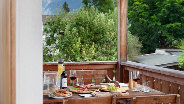 A wooden table set with food and drinks, a bottle of wine, and a scenic mountain view. - Falkenstein Apartment with Mountain View, Two Balconies, Private Sauna (Garmisch-Partenkirchen)