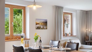 A dining area with a wooden table, chairs, a painting, and a window with curtains. - Falkenstein Apartment with Mountain View, Two Balconies, Private Sauna (Garmisch-Partenkirchen)