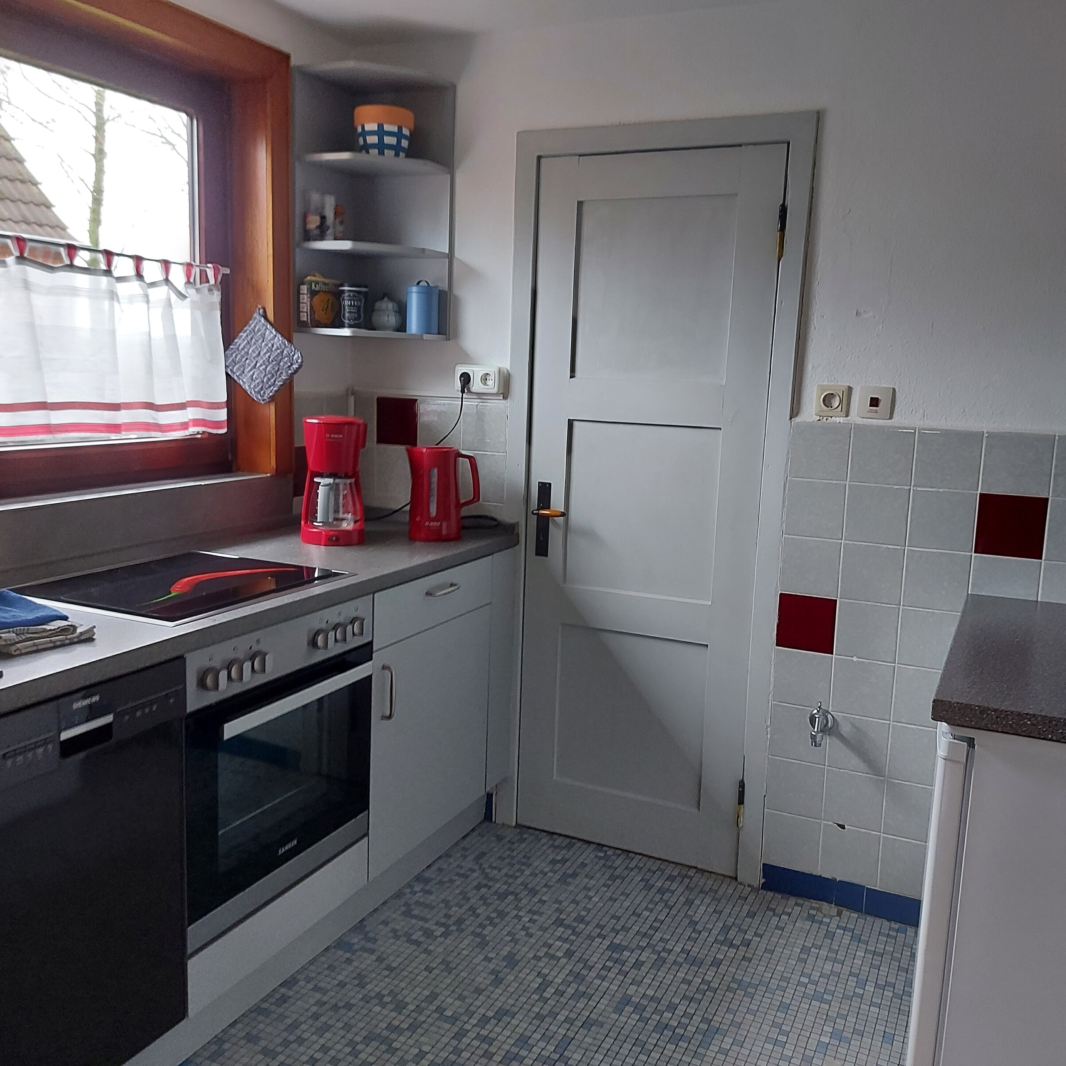 A kitchen with a dishwasher, oven, red coffee maker, and a window with curtains.