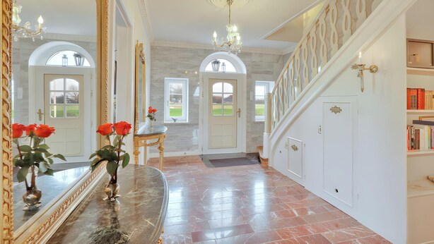 Un amplio pasillo con suelo de mármol, un espejo grande, una araña y una escalera con barandilla decorativa.