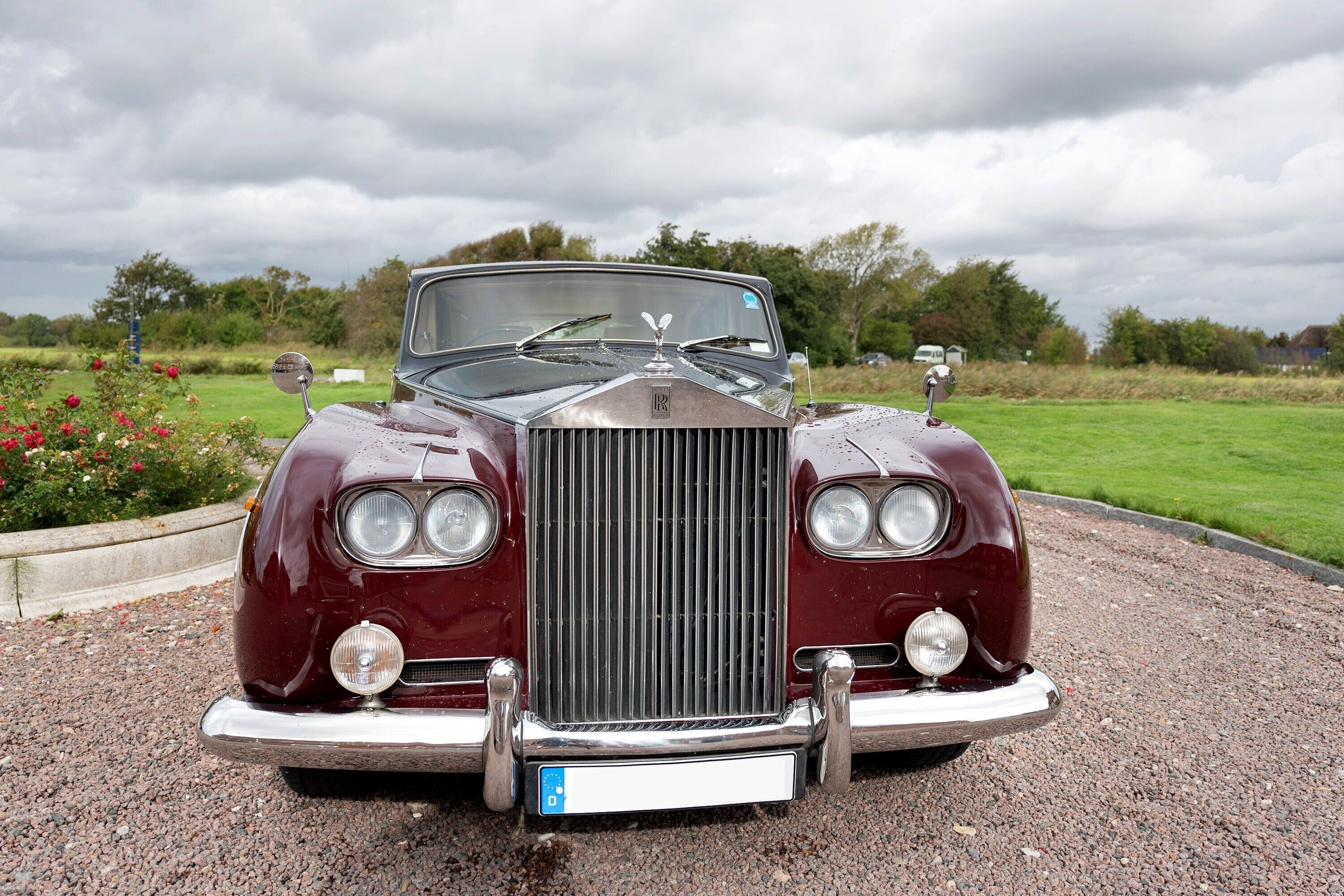 A classic car parked on a gravel path with a grassy field and trees in the background.