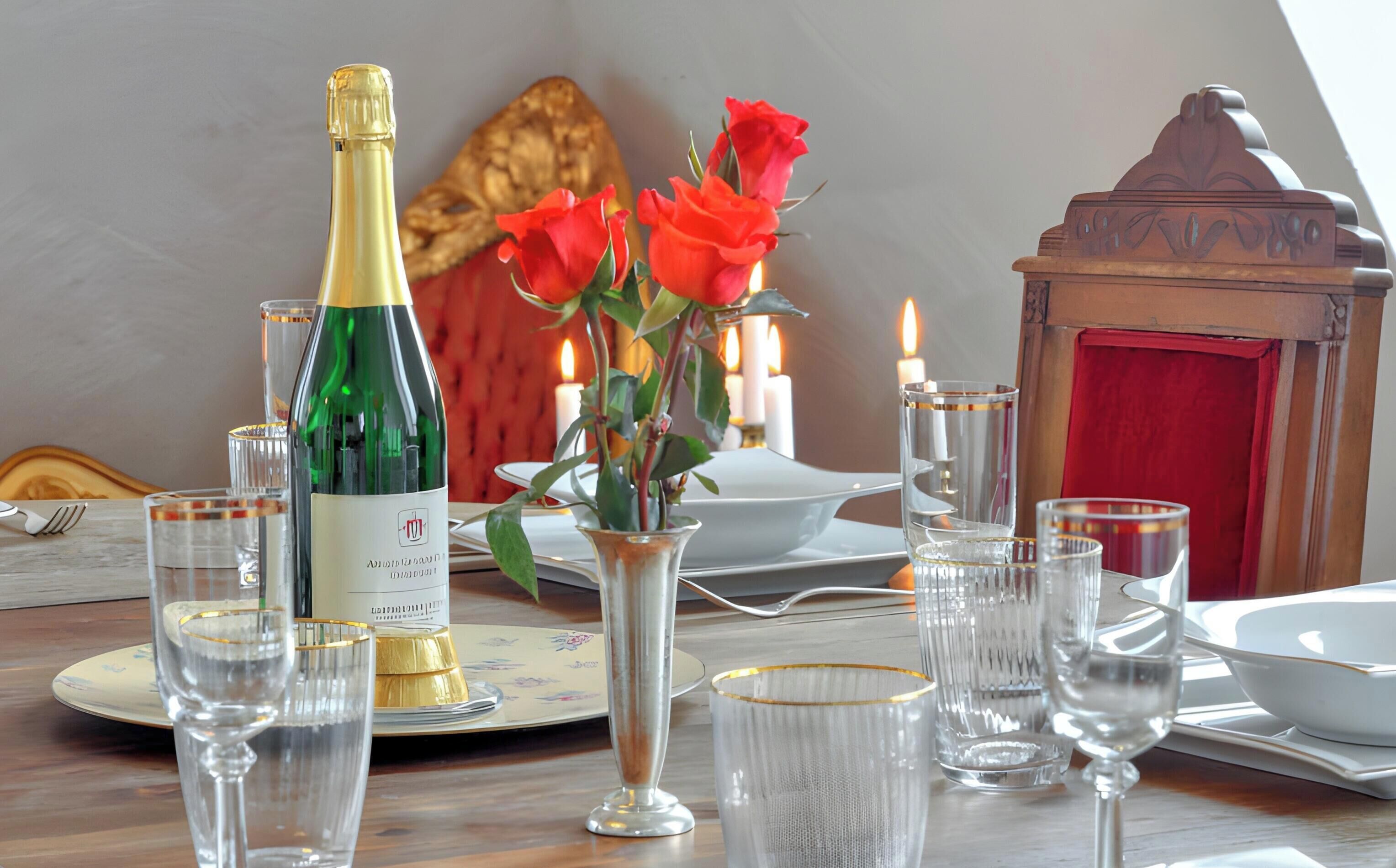 Ein Esstisch, gedeckt mit Champagnerflasche, Rosenvase und Kerzen im Hintergrund.