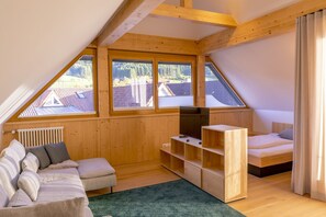 Ein gemütliches Dachzimmer mit schrägen Wänden, Holzwänden, einem fest installierten Fernseher, einer Couch, einem kleinen Bett und Blick auf Häuser durch große Fenster.