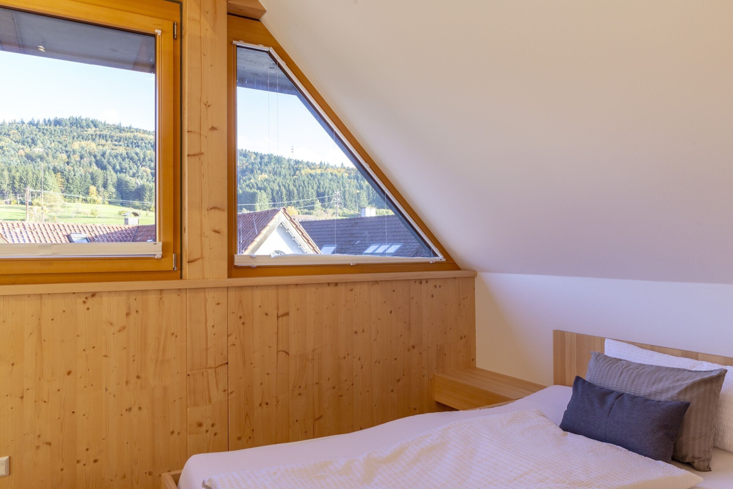 A bedroom with a sloped ceiling, wooden paneling, a bed with pillows, and a view of a forested area through large windows.
