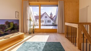 A room with a wooden floor, a TV on a wooden stand, a large window with curtains, and a view of buildings outside.