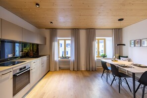 Une cuisine moderne dotée d’un four encastré, d’un coin repas avec une table et des chaises, et de grandes fenêtres avec des rideaux.