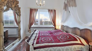 A bedroom with a large bed, ornate mirror, chandelier, and windows with curtains.