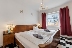 A hotel room with a checkered floor, two beds with white linens, a wooden headboard, a nightstand with a lamp, and a window with red curtains.