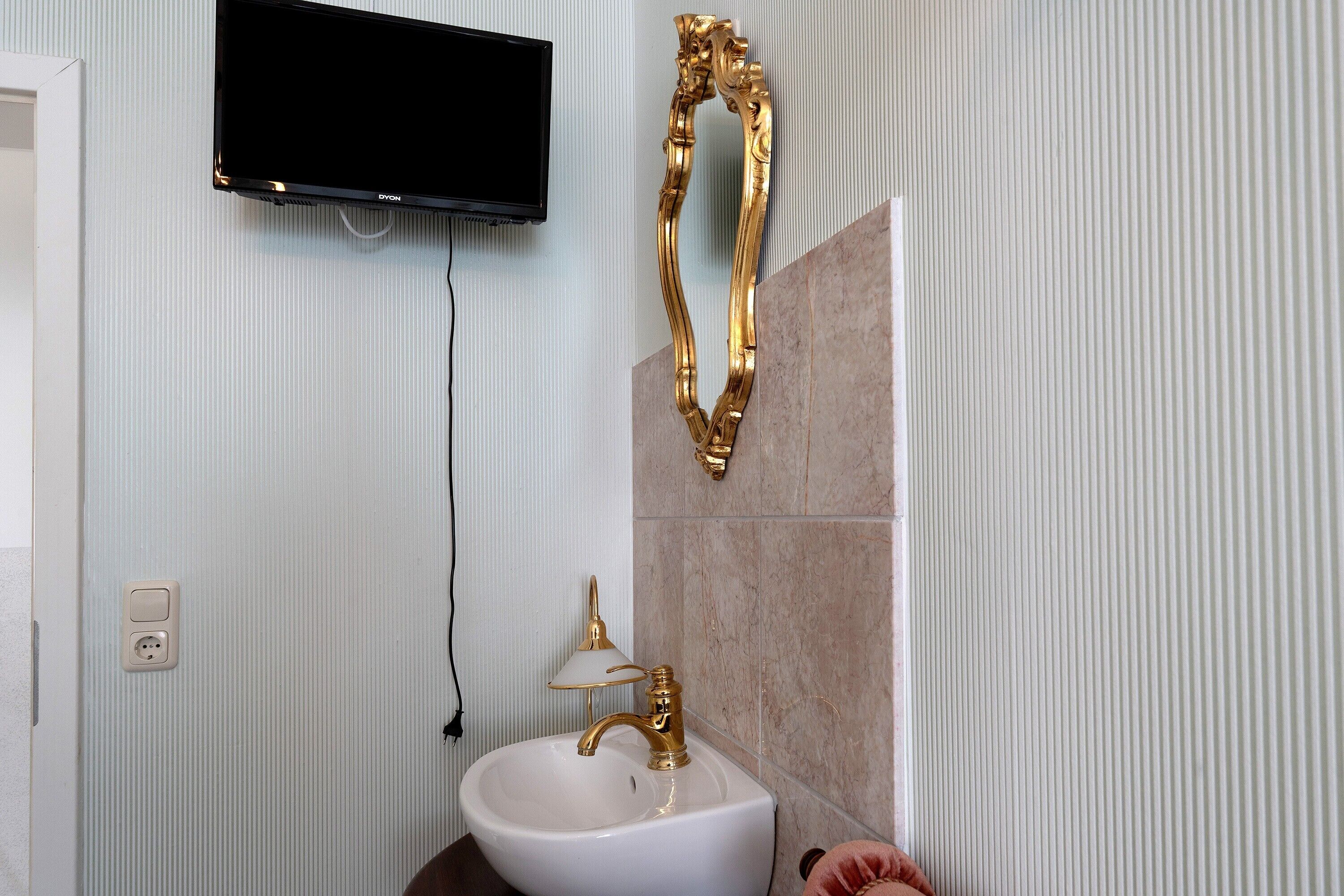 A bathroom with a white sink, a gold faucet, a mounted television, and a gold decorative piece.