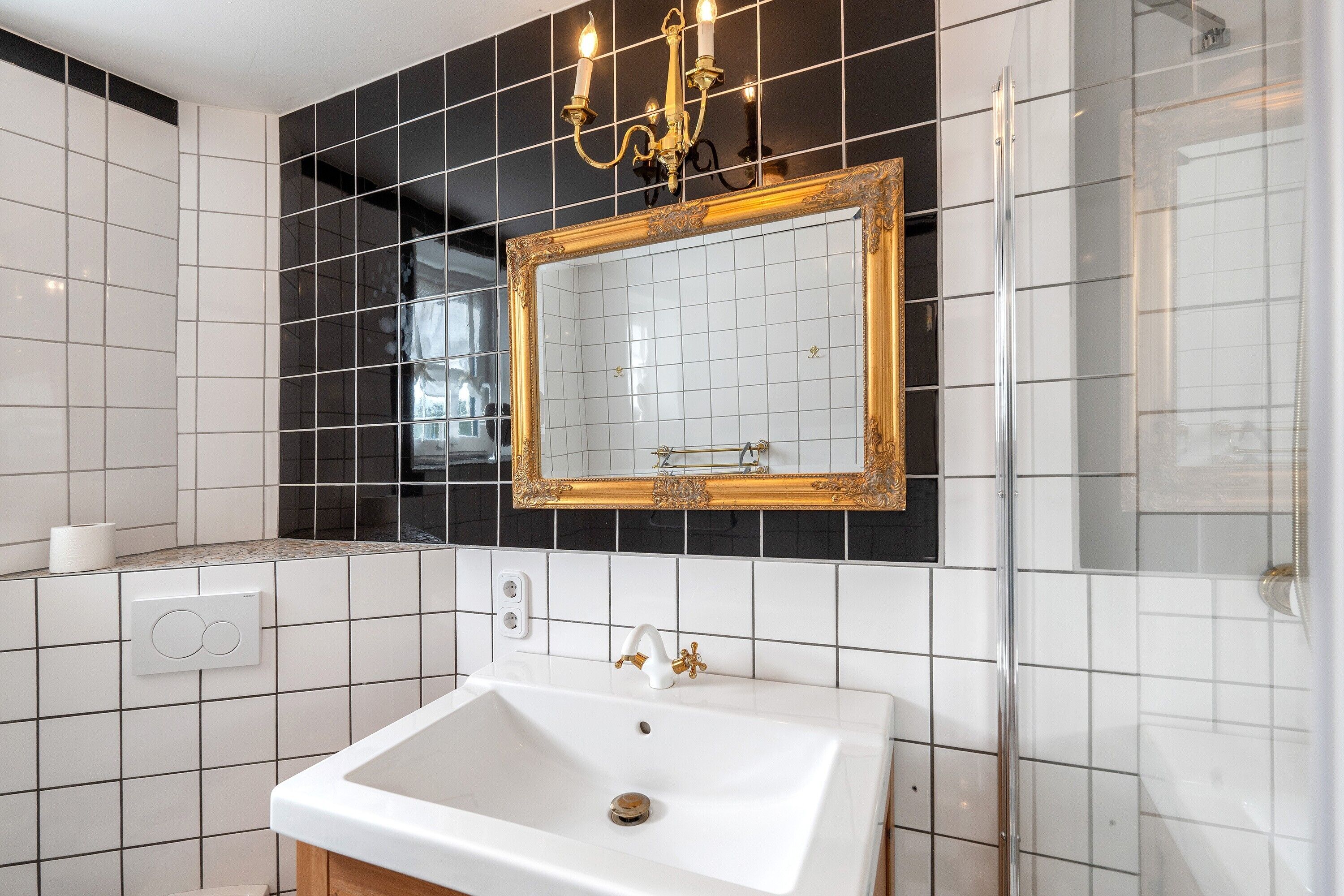 Ein Badezimmer mit einem weißen Waschbecken, einem spiegel mit goldener Rahmen und einer schwarzen Fliesenwand.