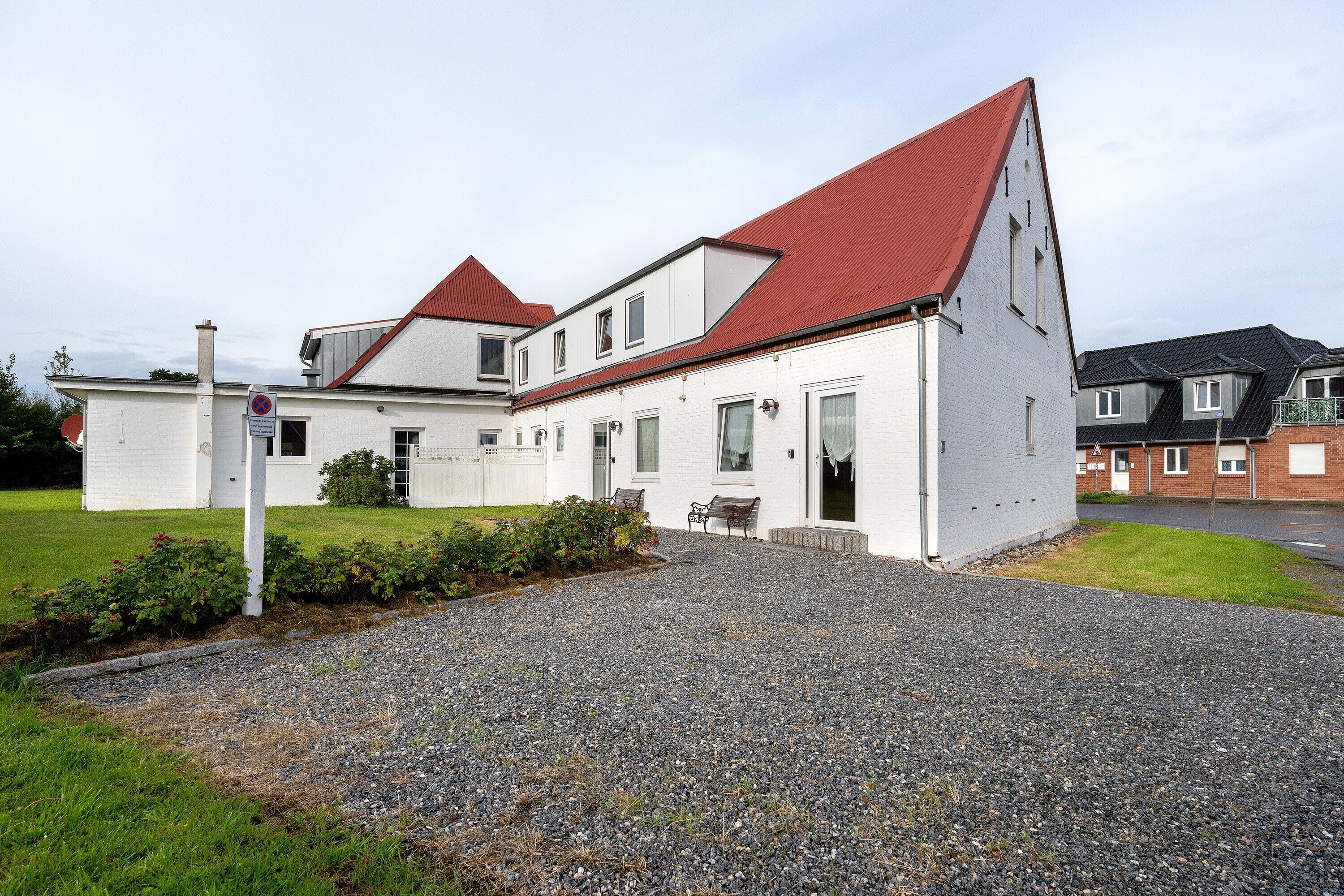 A white building with a red roof, a gravel driveway, and a small garden area.