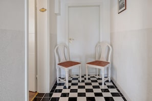A hallway with a checkered floor, two white chairs with brown cushions, a white door, and a small framed picture on the wall.