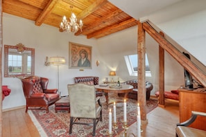 A room with a wooden ceiling, a chandelier, a red leather armchair, a white armchair, a round table with candles, and a painting on the wall.