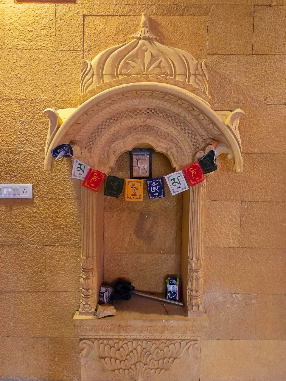 Un arco de piedra con un pequeño santuario en su interior, que incluye un cuadro enmarcado y una guirnalda de banderas de colores.