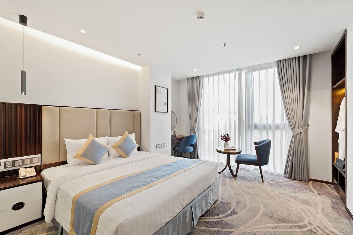 Une chambre d’hôtel avec un grand lit, une table de chevet, une lampe, une chaise et une vue sur la ville grâce à de grandes fenêtres.