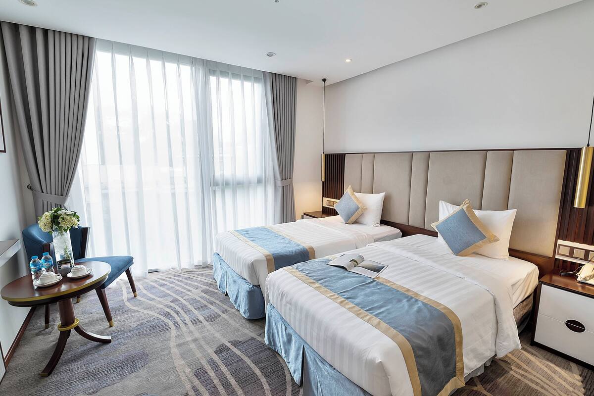 Une chambre d’hôtel avec deux lits, un bureau avec une chaise, un vase de fleurs et une grande fenêtre avec des rideaux.