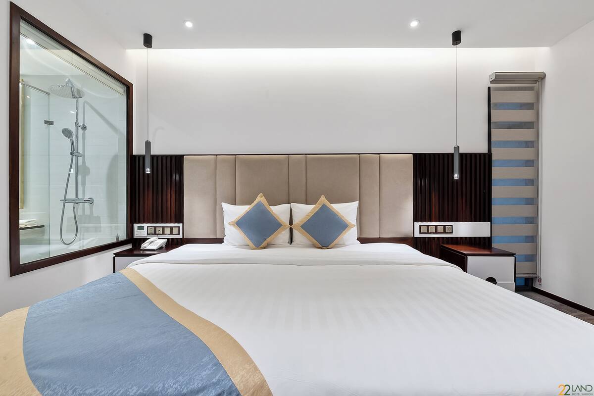 Une chambre d’hôtel moderne dotée d’un grand lit, d’une table de chevet avec un téléphone, d’une petite table de nuit, d’une cabine de douche en verre et offrant une vue sur la ville.