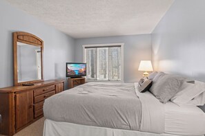 A bedroom with a bed, a dresser, a television, a window with blinds, and a lamp.
