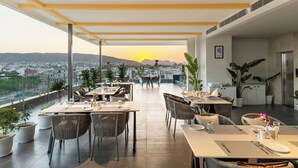 A modern dining area with tables and chairs, large potted plants, and a view of the city and mountains at sunset. - 7 Rays Hotel (Udaipur)