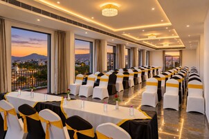 Un salón de conferencias con filas de mesas y sillas, ventanas amplias con vista a la ciudad y un balcón con vistas al atardecer.