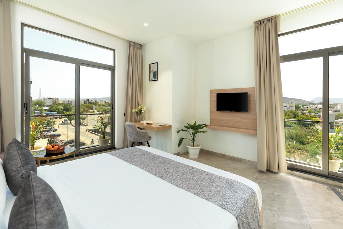 A hotel room with a large bed, a desk with a chair, a TV mounted on the wall, and a balcony with a view of the city.