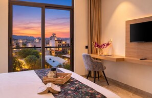 A hotel room with a large window, a bed, a desk with a chair, a TV, and a vase with flowers. - 7 Rays Hotel (Udaipur)