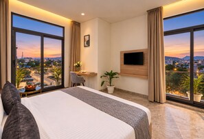 Ein Hotelzimmer mit einem großen Bett, einem Schreibtisch, einem Fernseher und Blick auf die Stadt bei Sonnenuntergang.