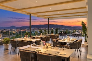 An outdoor dining area with tables and chairs, overlooking a cityscape at sunset. - 7 Rays Hotel (Udaipur)