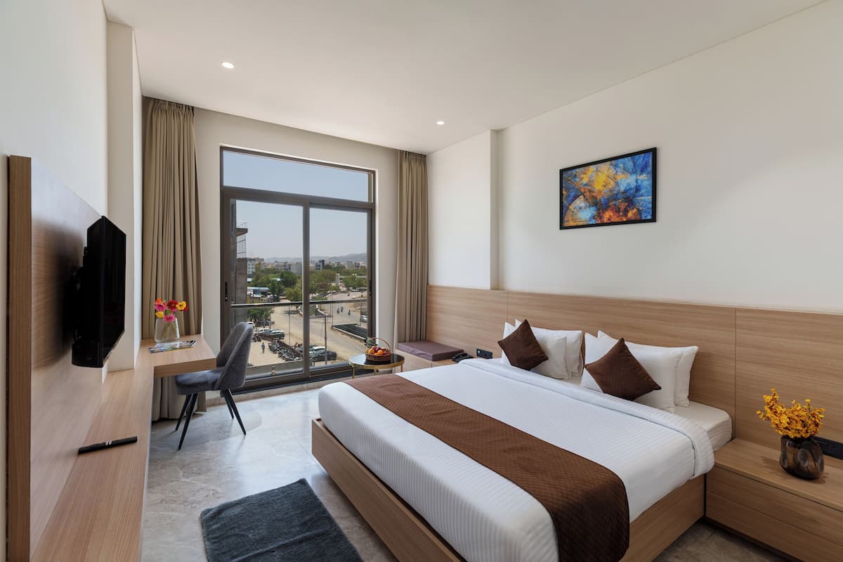 A modern hotel room with a large bed, a desk with a chair, a television, and a window with a city view.