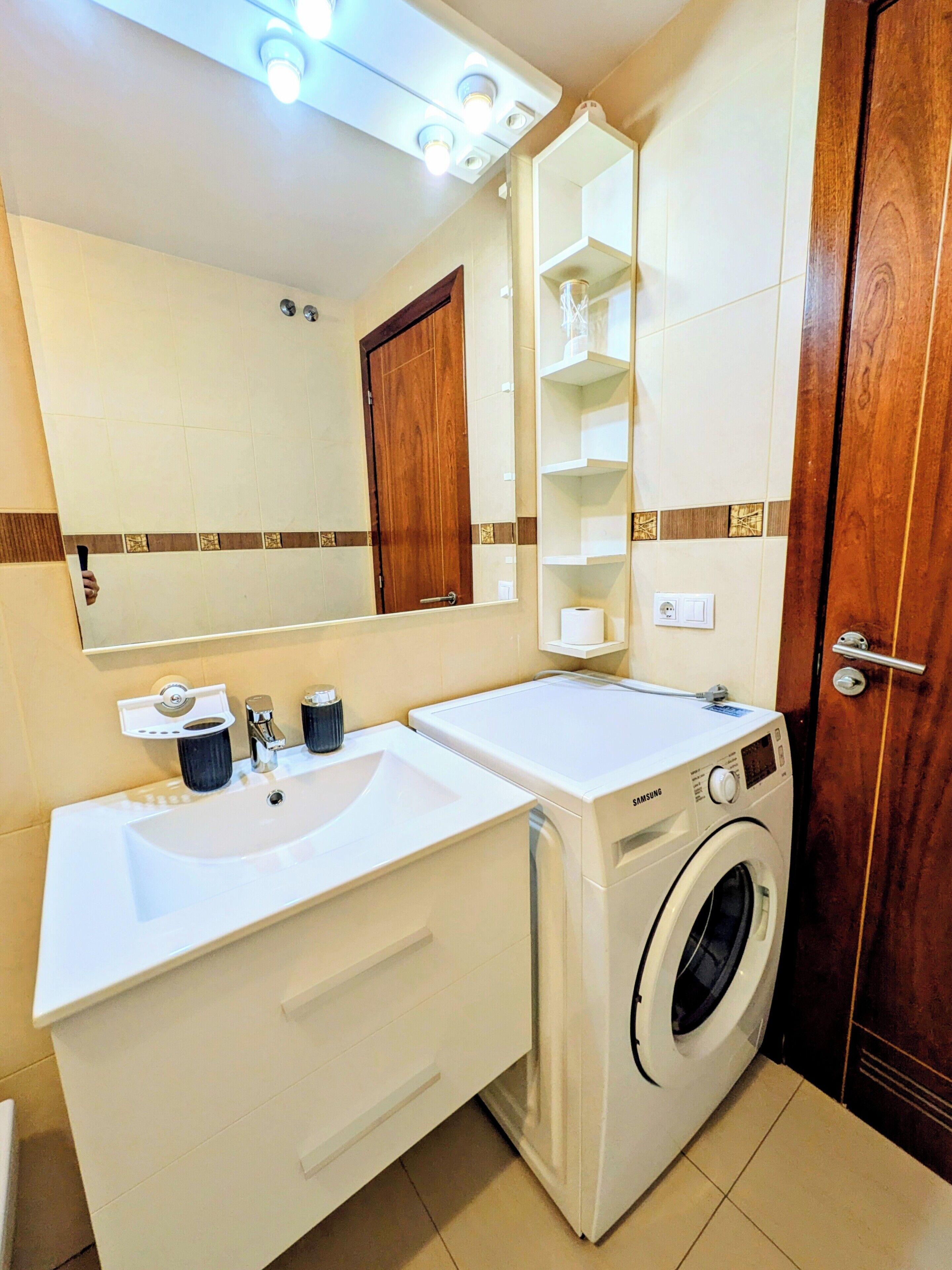 A bathroom with a double sink, a mirror, a washing machine, and a wooden door.