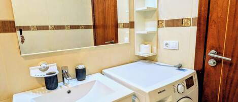 A bathroom with a double sink, a mirror, a washing machine, and a wooden door.