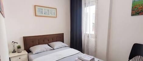 Un dormitorio con una cama, una mesita de noche, una silla, una ventana con cortinas y un cuadro en la pared.