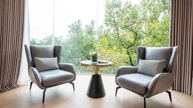 Una habitación moderna con dos sillones de cuero, una mesa redonda con un florero y un amplio ventanal que ofrece vistas a árboles y montañas.