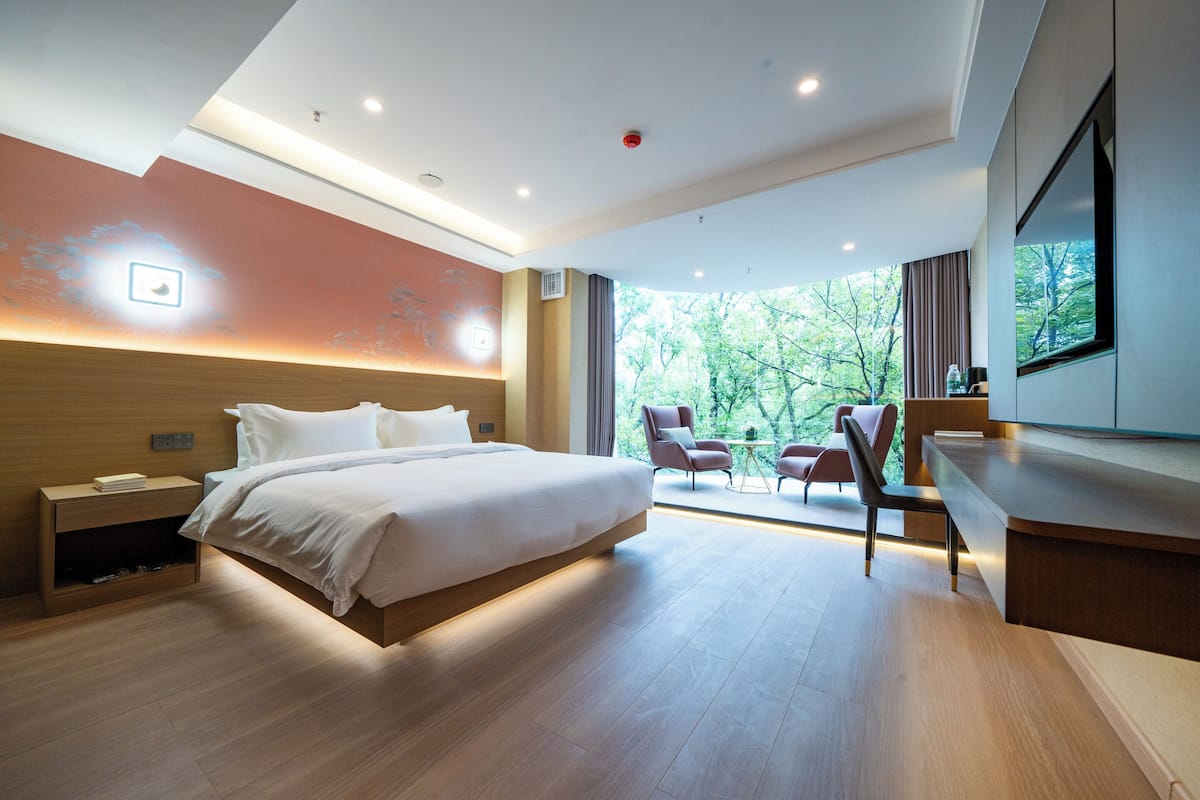 Moderni hotellihuone, jossa on suuri sänky, työpöytä ja ikkunasta avautuva näkymä puistoon.