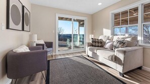 Un salón con un sofá y un sillón grises, y un tapete estampado. Hay dos ventanas amplias con vistas al exterior.