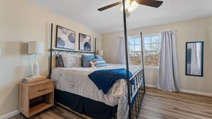 Un dormitorio con cama, mesita de noche, lámpara, ventilador de techo, ventana con cortinas y una obra de arte en la pared.
