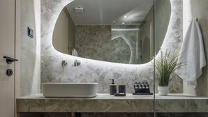 A modern bathroom with a large oval mirror, a built-in sink, and a shower area. - Loena Luxury Suites (Thessaloniki)