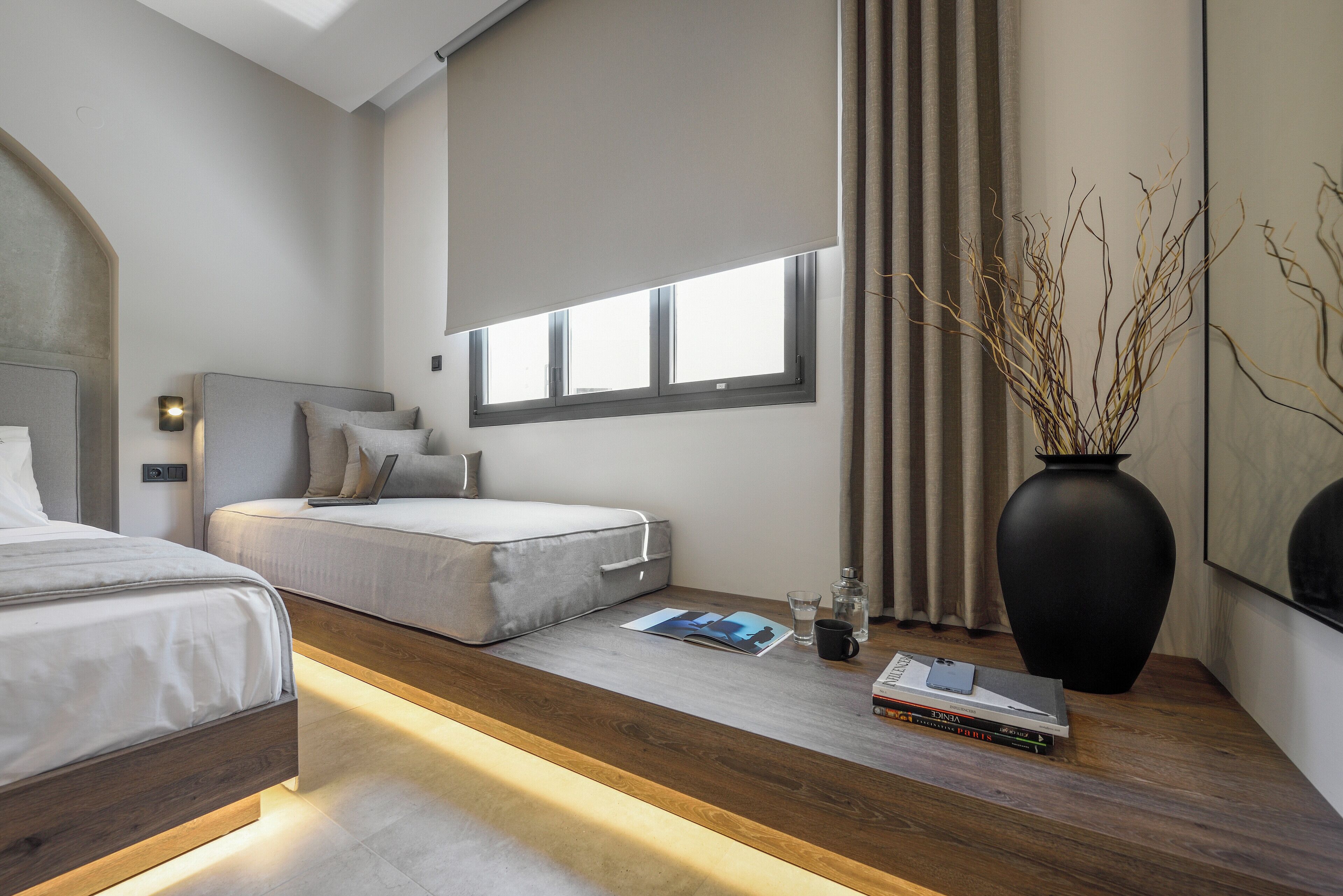 A modern bedroom with a wooden platform bed, a smaller bed, a bedside table with books and a vase, and a large window with curtains.